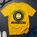 Beige t-shirt with 'Tunnel Champion Agility Handler' graphic on a dark surface with shoes and jeans.