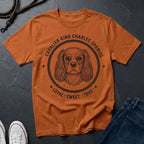 Orange Comfort Colors t-shirt with Cavalier King Charles Spaniel graphic and text on a dark surface.