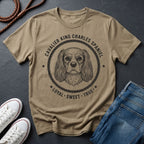 Beige Comfort Colors t-shirt with Cavalier King Charles Spaniel graphic and text on a dark surface with shoes and jeans.