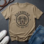 Beige Comfort Colors t-shirt with dachshund graphic and text on a dark surface with shoes and jeans.