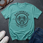 Teal Comfort Colors t-shirt with dachshund graphic and text on a dark surface with shoes and jeans.