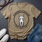 Beige Comfort Colors t-shirt with Doberman Pinscher graphic and text on a dark surface with shoes and jeans.