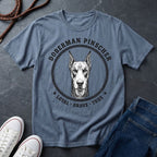 Blue Comfort Colors t-shirt with Doberman Pinscher graphic and text on a dark surface with shoes and jeans.