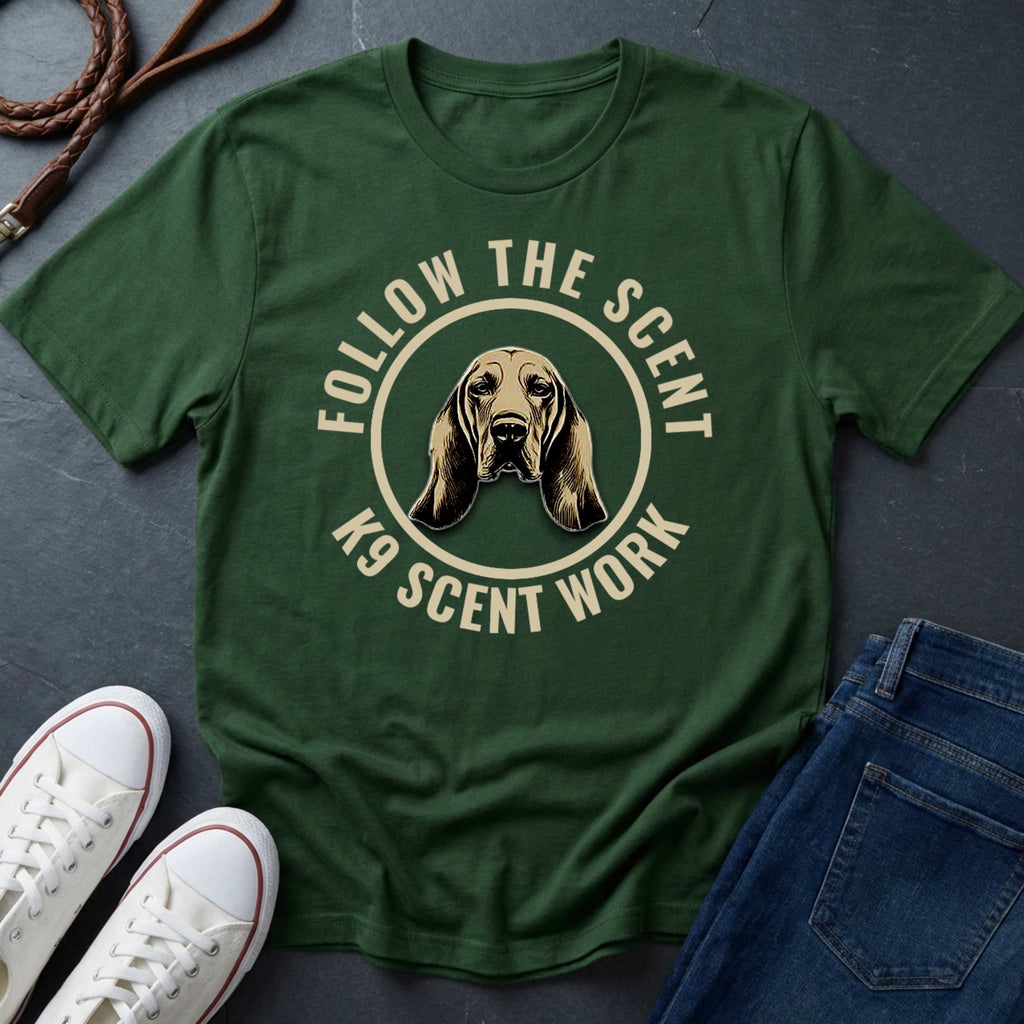 Green Comfort Colors t-shirt with a dog graphic and text 'Follow the Scent K9 Scent Work' on a dark surface with shoes and jeans.