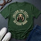 Green Comfort Colors t-shirt with a dog graphic and text 'Follow the Scent K9 Scent Work' on a dark surface with shoes and jeans.