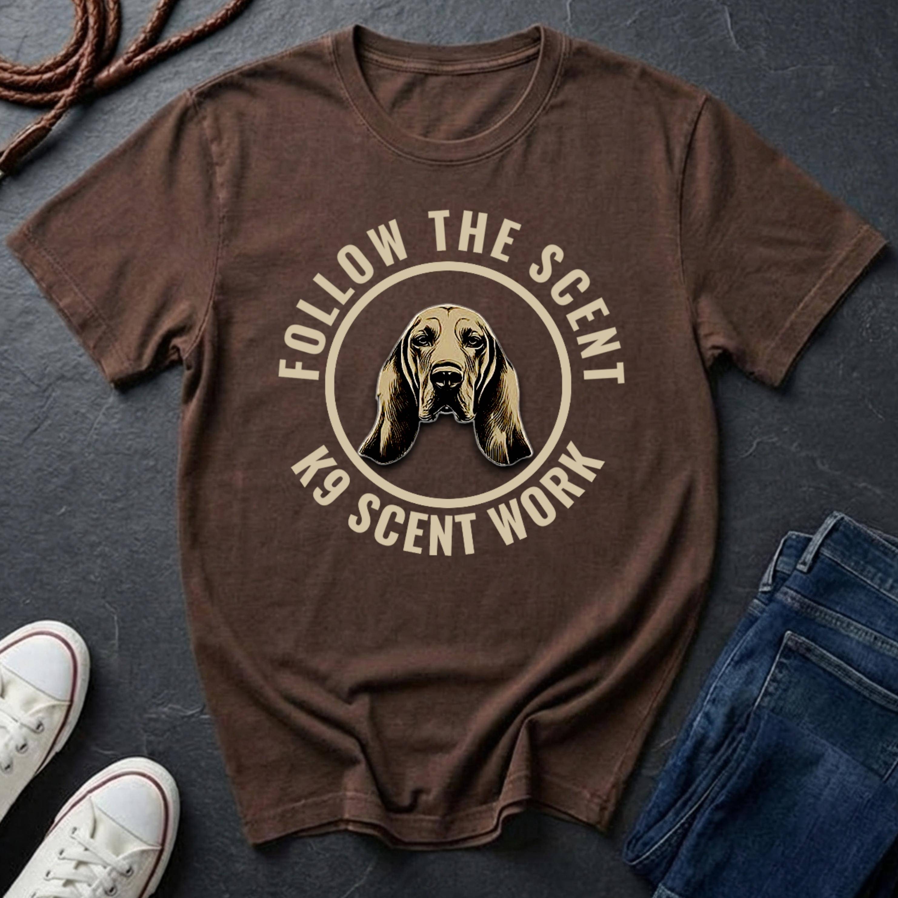 Brown Comfort Colors t-shirt with dog graphic and text 'Follow the Scent K9 Scent Work' on a dark surface with shoes and jeans.