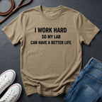 Beige Comfort Colors t-shirt with text 'I work hard so my lab can have a better life' on a dark surface with shoes and jeans.