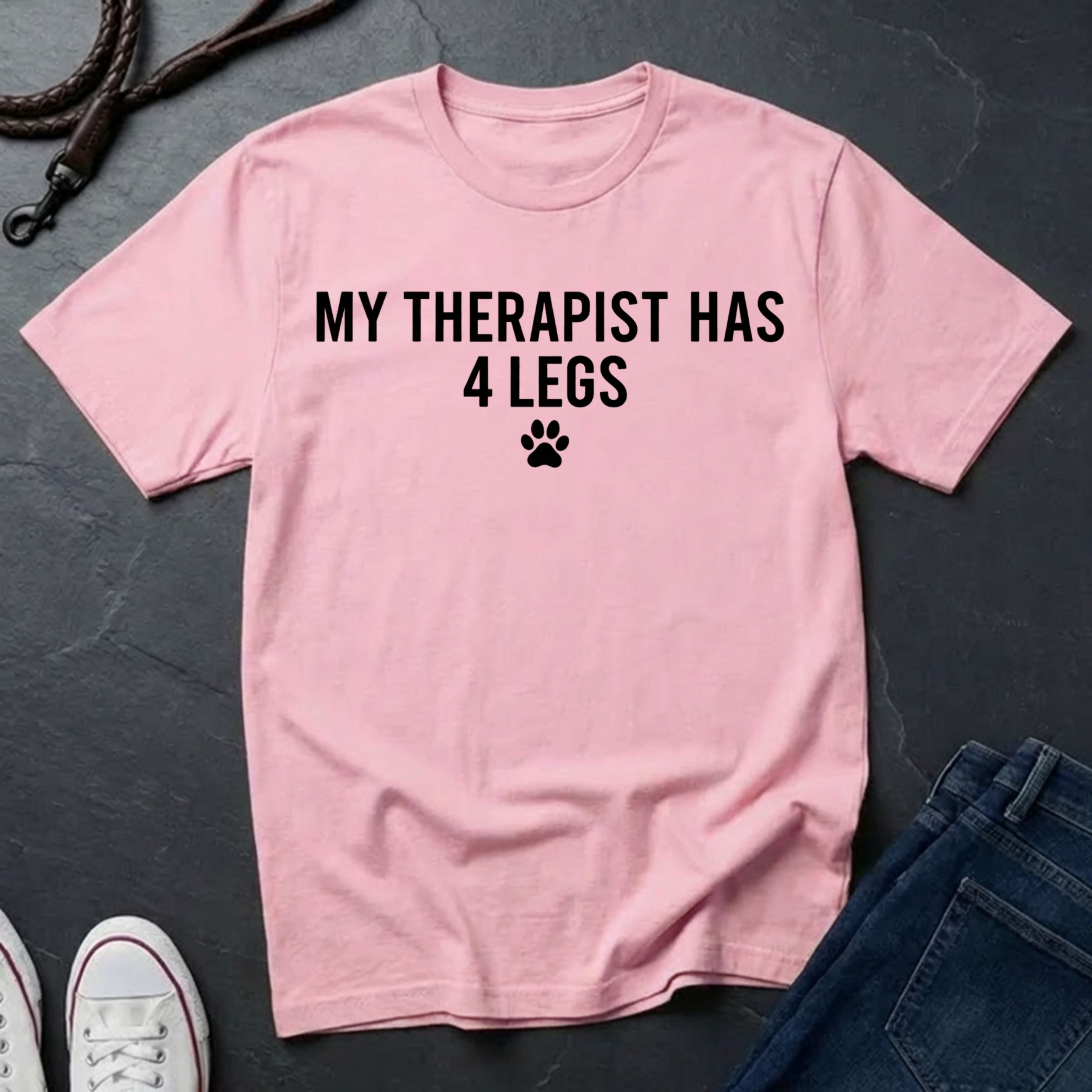 Pink Comfort Colors t-shirt with 'My therapist has 4 legs' text on a dark surface with jeans and shoes.