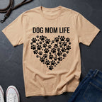 Beige Comfort Colors t-shirt with 'DOG MOM LIFE' text and paw print heart design on a dark surface.