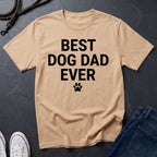 Beige Comfort Colors t-shirt with 'Best Dog Dad Ever' text on a dark surface with jeans and shoes.
