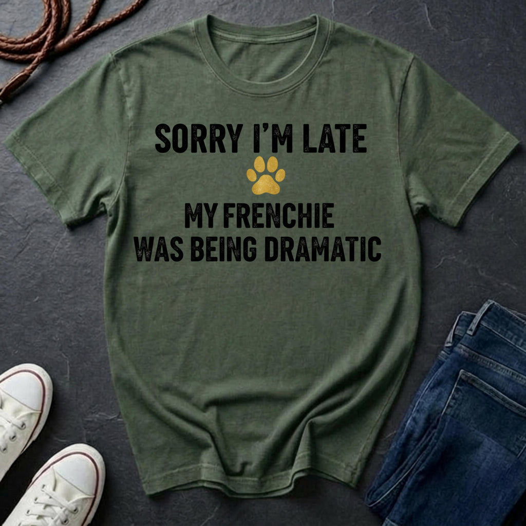 Green Comfort Colors t-shirt with text 'Sorry I'm late, my Frenchie was being dramatic' on a dark surface with shoes and jeans.