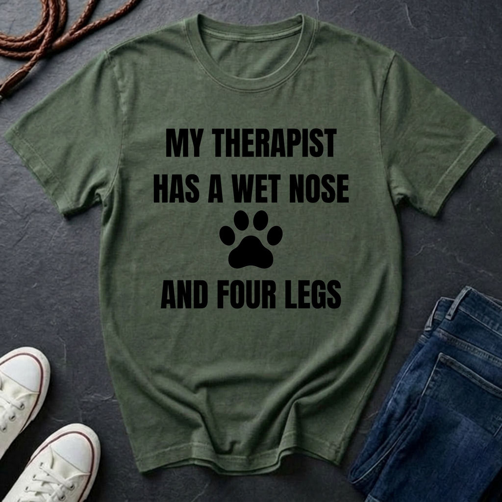 Green Comfort Colors t-shirt with text 'My therapist has a wet nose and four legs' on a dark surface with jeans and shoes.