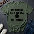 Green Comfort Colors t-shirt with text 'My therapist has a wet nose and four legs' on a dark surface with jeans and shoes.