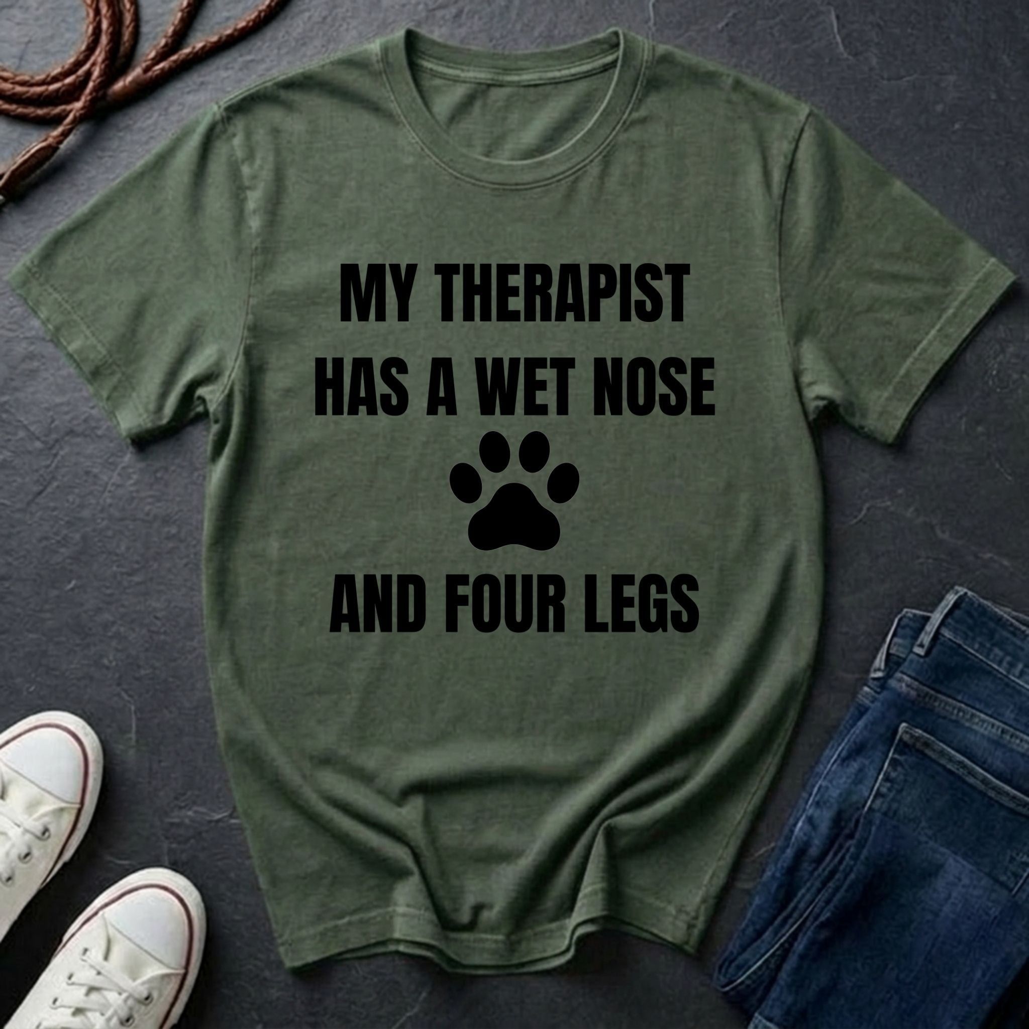 Green Comfort Colors t-shirt with text 'My therapist has a wet nose and four legs' on a dark surface with jeans and shoes.