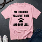 Pink Comfort Colors t-shirt with text 'My therapist has a wet nose and four legs' on a dark surface.