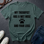 Green Comfort Colors t-shirt with text 'My therapist has a wet nose and four legs' on a dark surface with shoes and jeans.