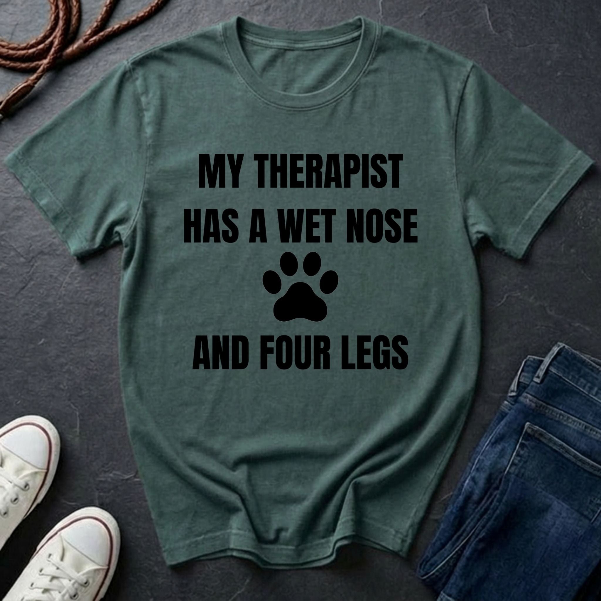 Green Comfort Colors t-shirt with text 'My therapist has a wet nose and four legs' on a dark surface with shoes and jeans.