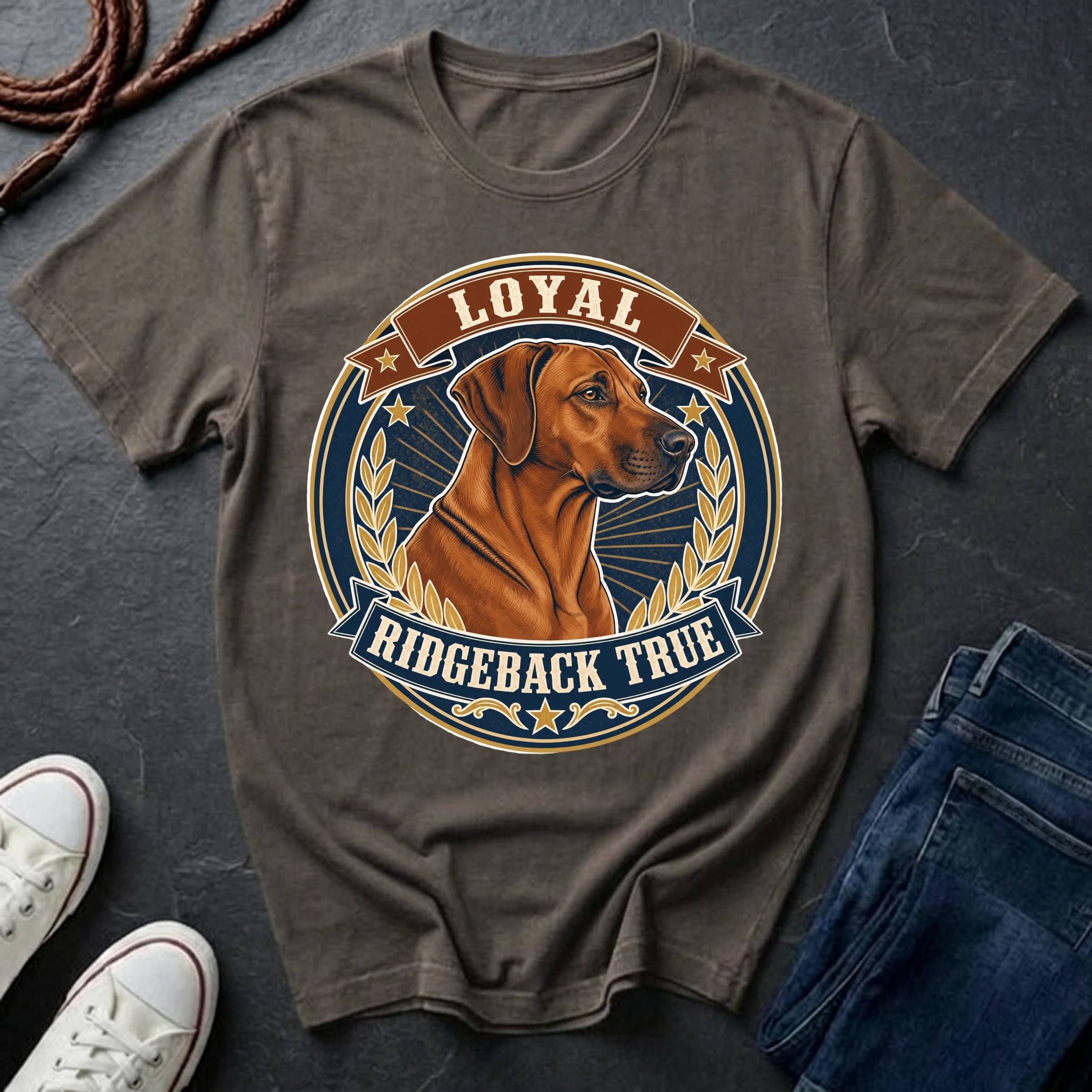 A pepper graphic t-shirt featuring a Rhodesian Ridgeback dog illustration with the text 'LOYAL RIDGEBACK TRUE' in a circular badge design. The t-shirt is styled in a flat-lay arrangement on a dark slate surface, accompanied by white sneakers, a coiled rope, and a pair of blue jeans.