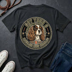 A black graphic t-shirt featuring a Cavalier King Charles Spaniel dog illustration inside a circular badge design, styled in a flat lay with jeans, sneakers, and a leather strap on a dark slate background.