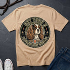 A Ivory short-sleeve t-shirt laid flat on a dark slate surface, featuring a large circular graphic of a Cavalier King Charles Spaniel dog with the text 'GOT YOUR 6 CAVALIER KING CHARLES 6'. White sneakers and blue jeans are partially visible as styling props.