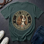 A blue spruce graphic t-shirt laid flat on a dark slate background, styled with white sneakers, a coiled rope, and a pair of blue jeans. The back of the t-shirt features a large circular graphic of a Cavalier King Charles Spaniel dog inside a distressed emblem with the text 'GOT YOUR 6 CANVASIER KING CHARLES 6'.