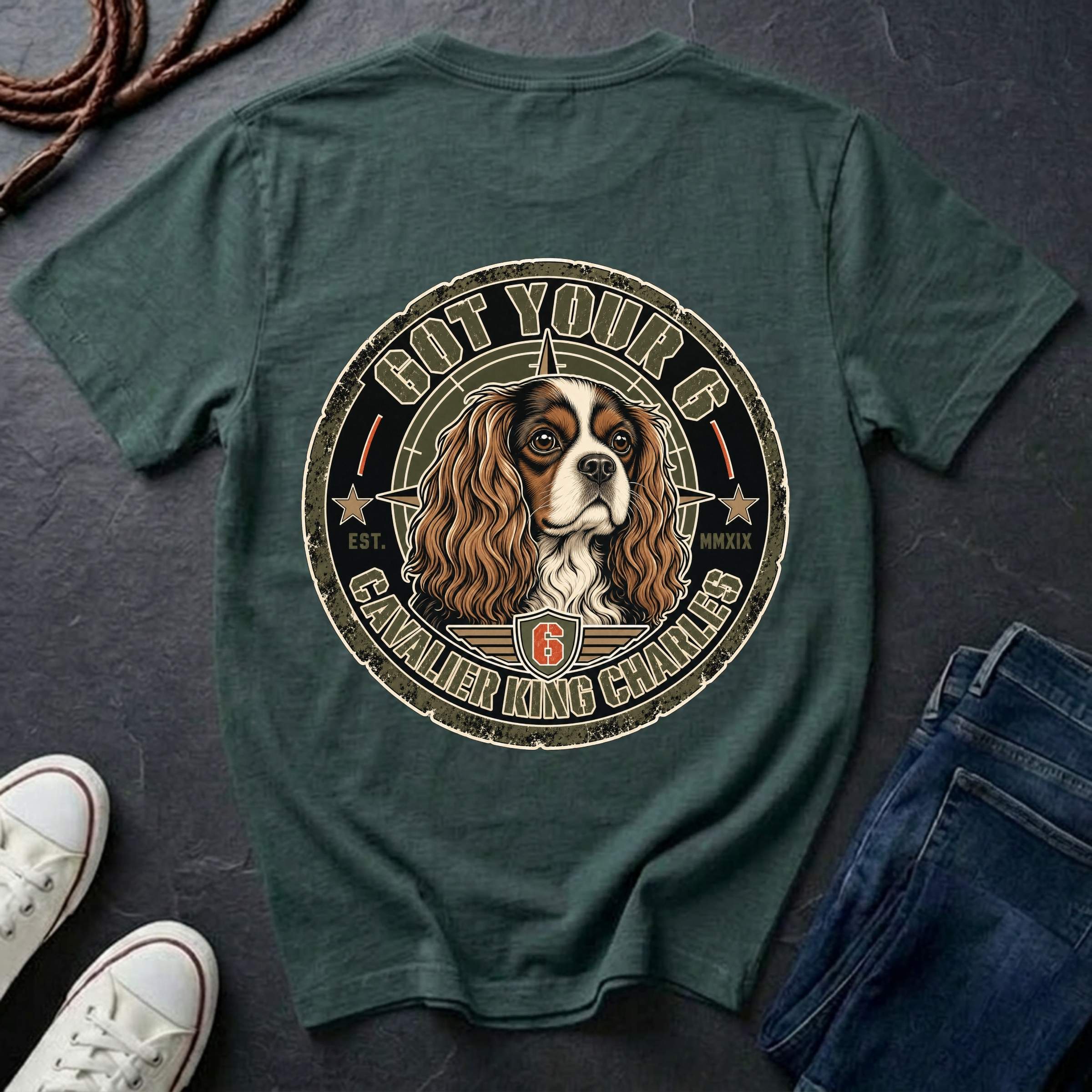 A blue spruce graphic t-shirt laid flat on a dark slate background, styled with white sneakers, a coiled rope, and a pair of blue jeans. The back of the t-shirt features a large circular graphic of a Cavalier King Charles Spaniel dog inside a distressed emblem with the text 'GOT YOUR 6 CANVASIER KING CHARLES 6'.