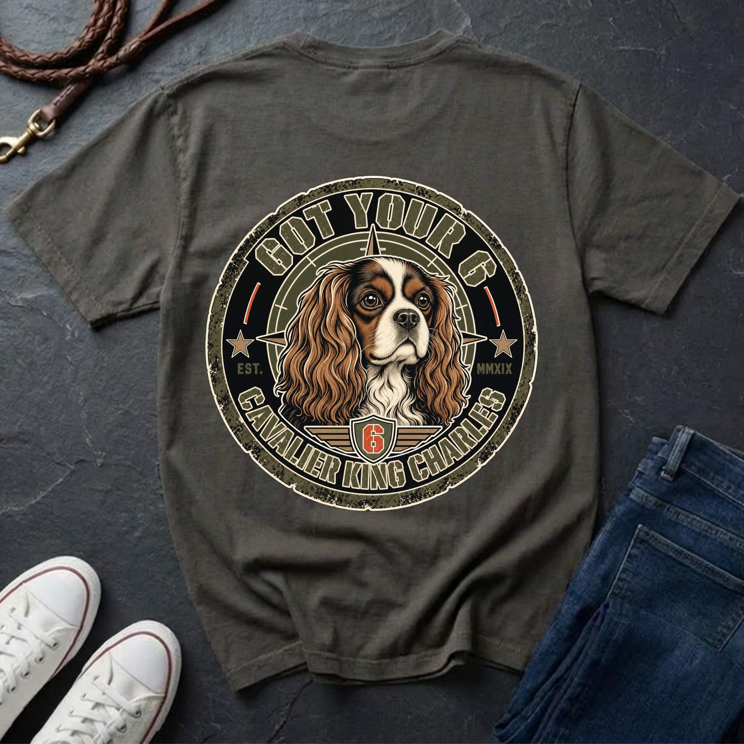 A dark pepper t-shirt laid flat on a dark slate background, styled with white sneakers, blue jeans, and a leather strap. The back of the shirt features a large circular graphic of a Cavalier King Charles Spaniel dog inside a seal with the text 'GOT YOUR 6 CAVALIER KING CHARLES 6'.