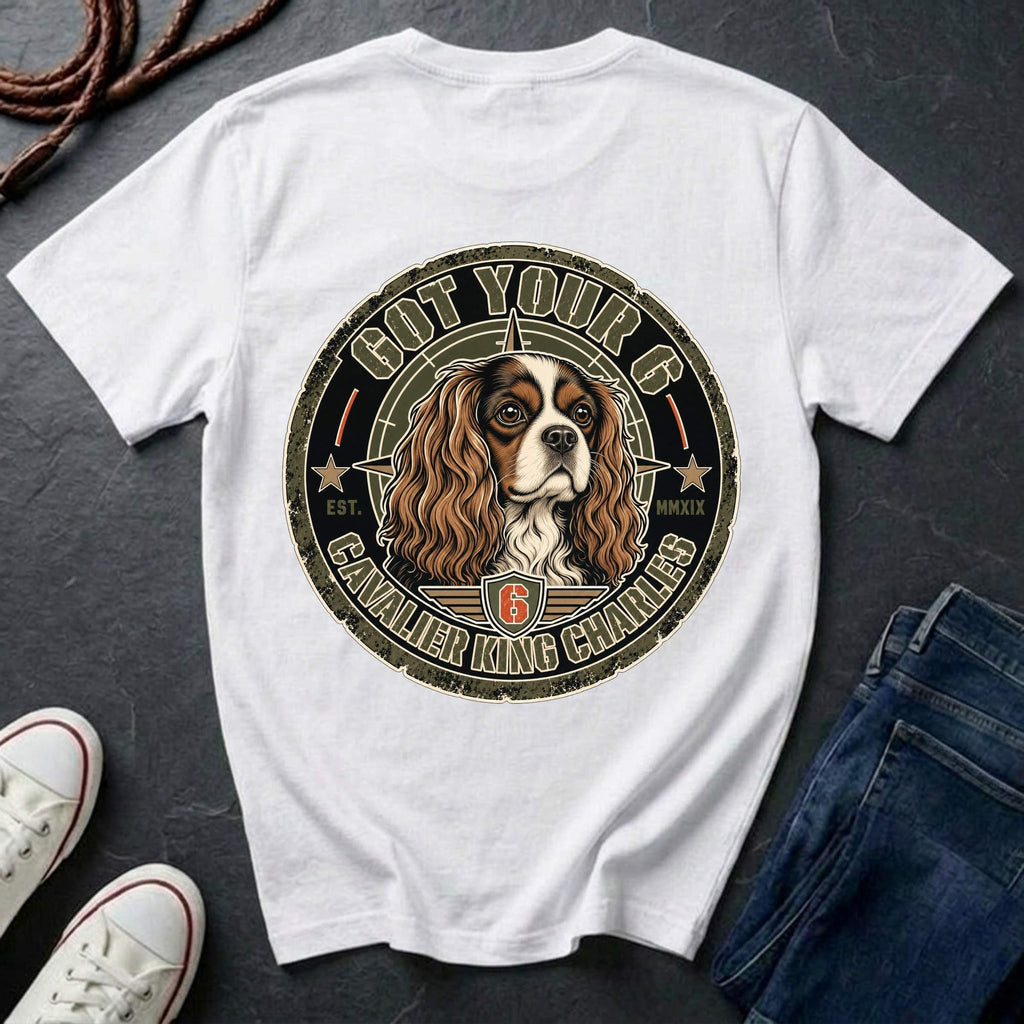 A white short-sleeve T-shirt laid flat on a dark textured surface, featuring a large circular graphic of a Cavalier King Charles Spaniel dog with the text 'GOT YOUR 6' and 'CAVALIER KING CHARLES' around it. Jeans, sneakers, and rope are styled around the shirt.