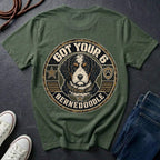 An bay short-sleeve t-shirt laid flat on a dark slate surface, featuring a large circular graphic of a Bernedoodle dog with the text 'GOT YOUR 6 BERNEDOOLE' and a paw print. White sneakers and blue jeans are partially visible as styling props.