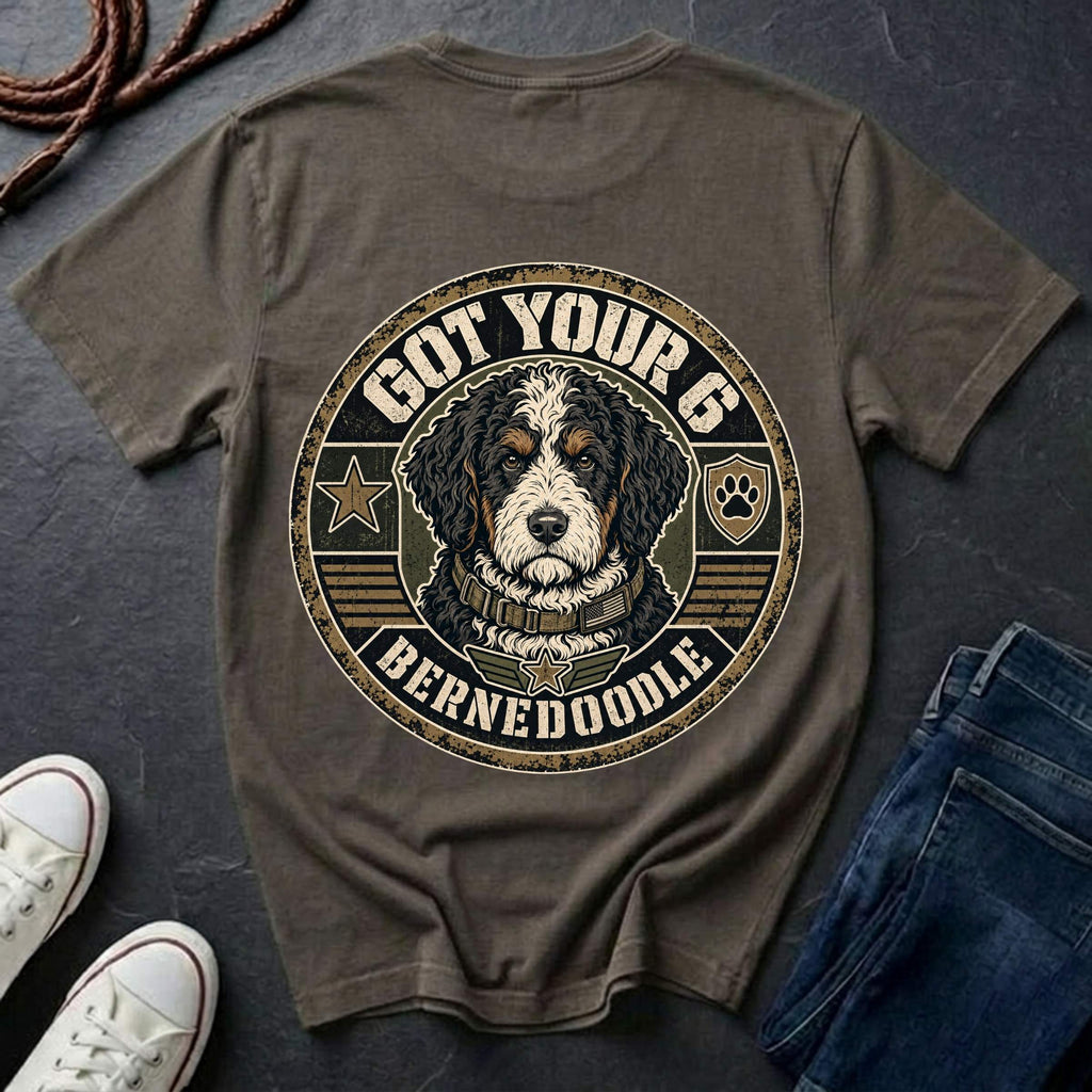 A pepper t-shirt with a large circular graphic of a Bernedoodle dog and the text 'GOT YOUR 6 BERNEDOOLE' on the back, styled in a flat lay with jeans, sneakers, and a rope on a dark slate background.