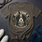 A pepper t-shirt with a large circular graphic of a Bernedoodle dog and the text 'GOT YOUR 6 BERNEDOOLE' on the back, styled in a flat lay with jeans, sneakers, and a rope on a dark slate background.