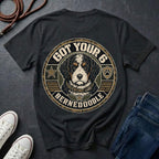 A black graphic t-shirt featuring a Bernedoodle dog illustration inside a circular badge design with the text 'GOT YOUR 6 BERNEDOODLE'. The shirt is styled in a flat-lay arrangement on a dark slate background with jeans, sneakers, and a rope.
