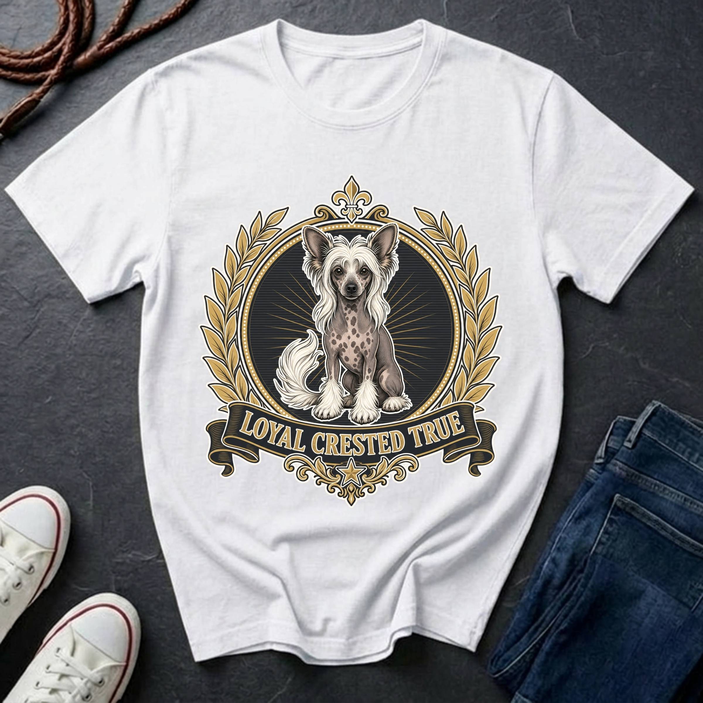 A white short-sleeve graphic T-shirt laid flat on a dark textured surface, featuring a decorative crest with a Chinese Crested dog illustration and the text 'LOYAL CRESTED TRUE.' Blue jeans and white sneakers are partially visible around it as styling props.