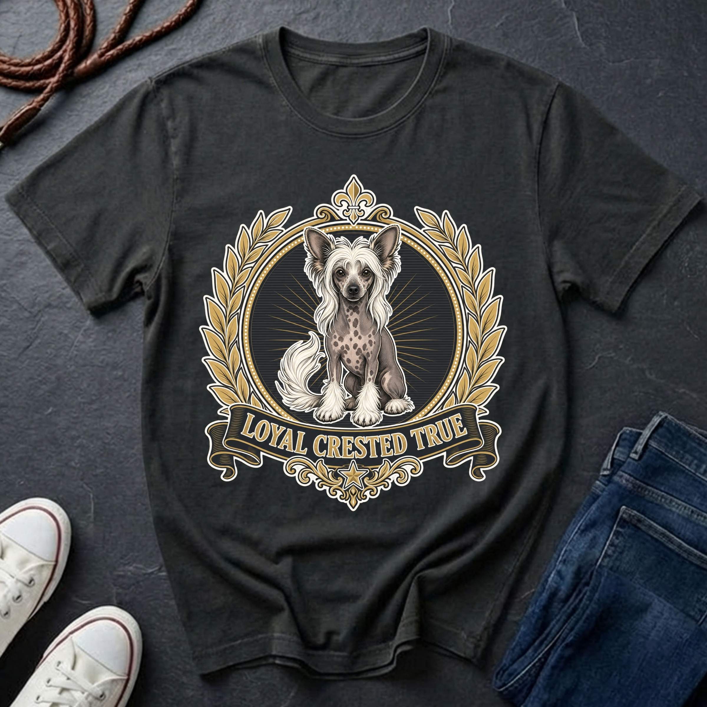 A black graphic t-shirt featuring a detailed illustration of a Chinese Crested dog inside an ornate gold and beige crest with the text 'LOYAL CRESTED TRUE'. The shirt is displayed in a flat-lay arrangement on a dark slate background, styled with white sneakers, a coiled rope, and blue jeans.