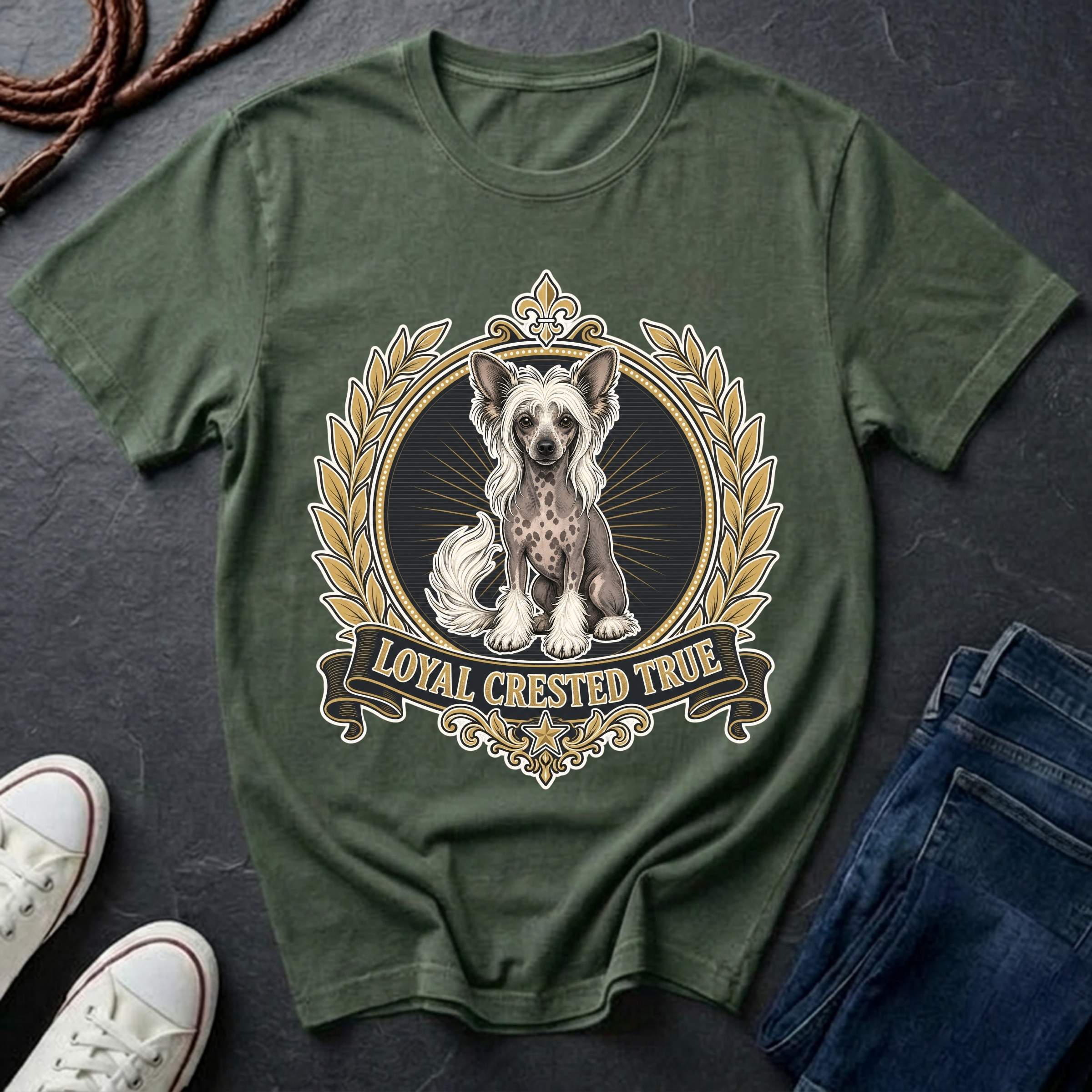 A bay short-sleeve t-shirt laid flat on a dark slate background, featuring a detailed graphic of a Chinese Crested dog inside a gold laurel wreath with the text 'LOYAL CRESTED TRUE'. White sneakers and blue jeans are partially visible as styling props.