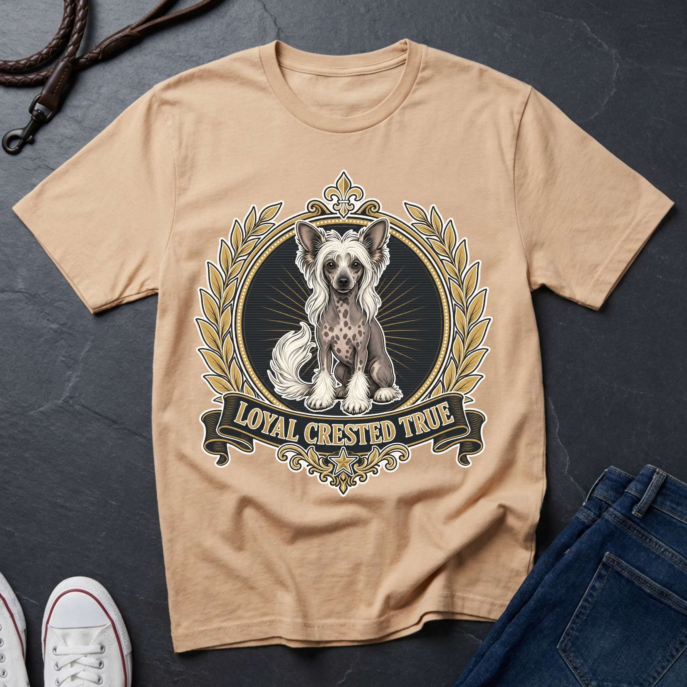 A ivory crewneck t-shirt laid flat on a dark slate background, featuring a graphic of a Chinese Crested dog inside an ornate gold and black circular frame with the text 'LOYAL CRESTED TRUE'. White sneakers and blue jeans are partially visible as styling props.