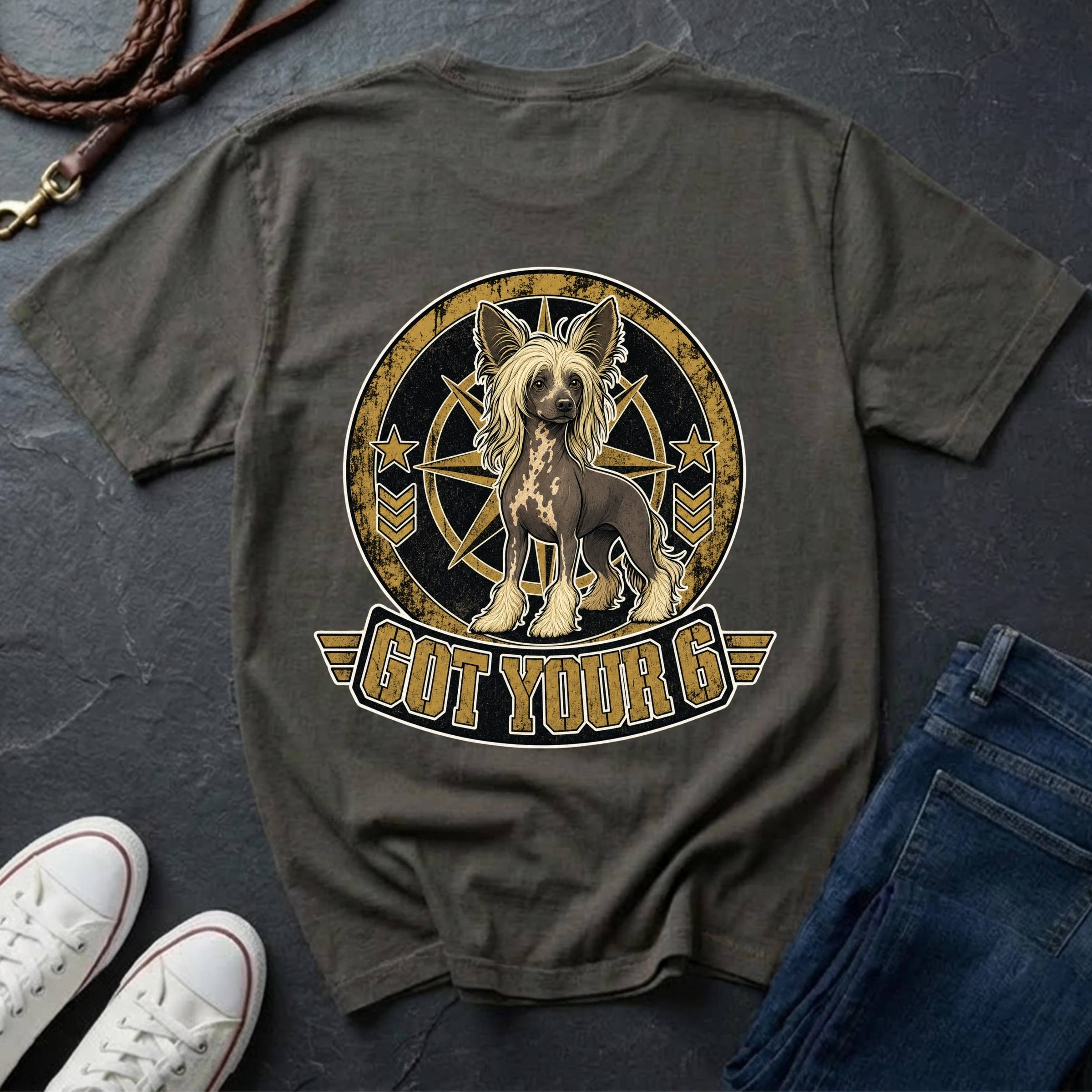 A grey t-shirt laid flat on a dark slate surface, featuring a graphic of a Chinese Crested dog inside a circular emblem with the text 'GOT YOUR 6'. White sneakers and blue jeans are styled around it with a leather strap in the corner.