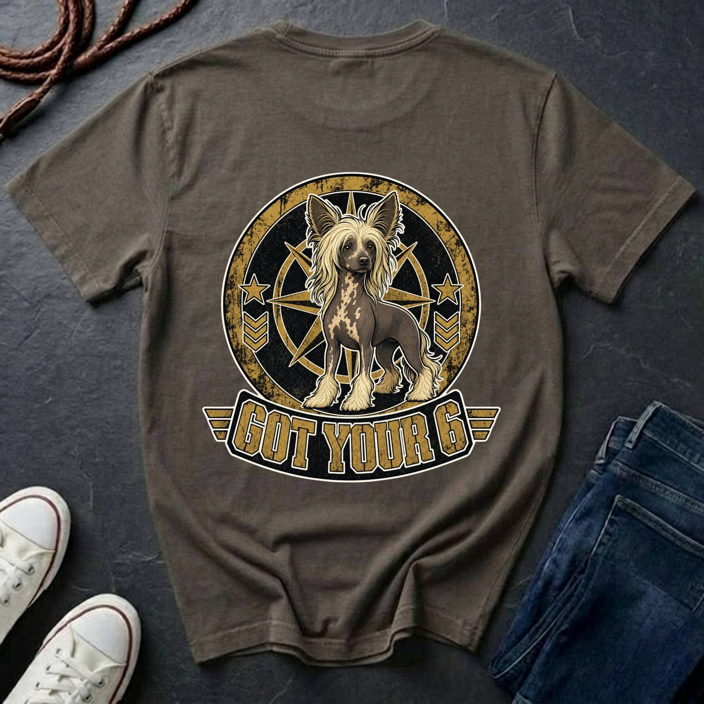 A dark brown t-shirt featuring a graphic of a Chinese Crested dog inside a circular emblem with the text 'GOT YOUR 6' below it. The shirt is styled in a flat-lay arrangement on a dark slate background, accompanied by white sneakers, a coiled rope, and a pair of blue jeans.